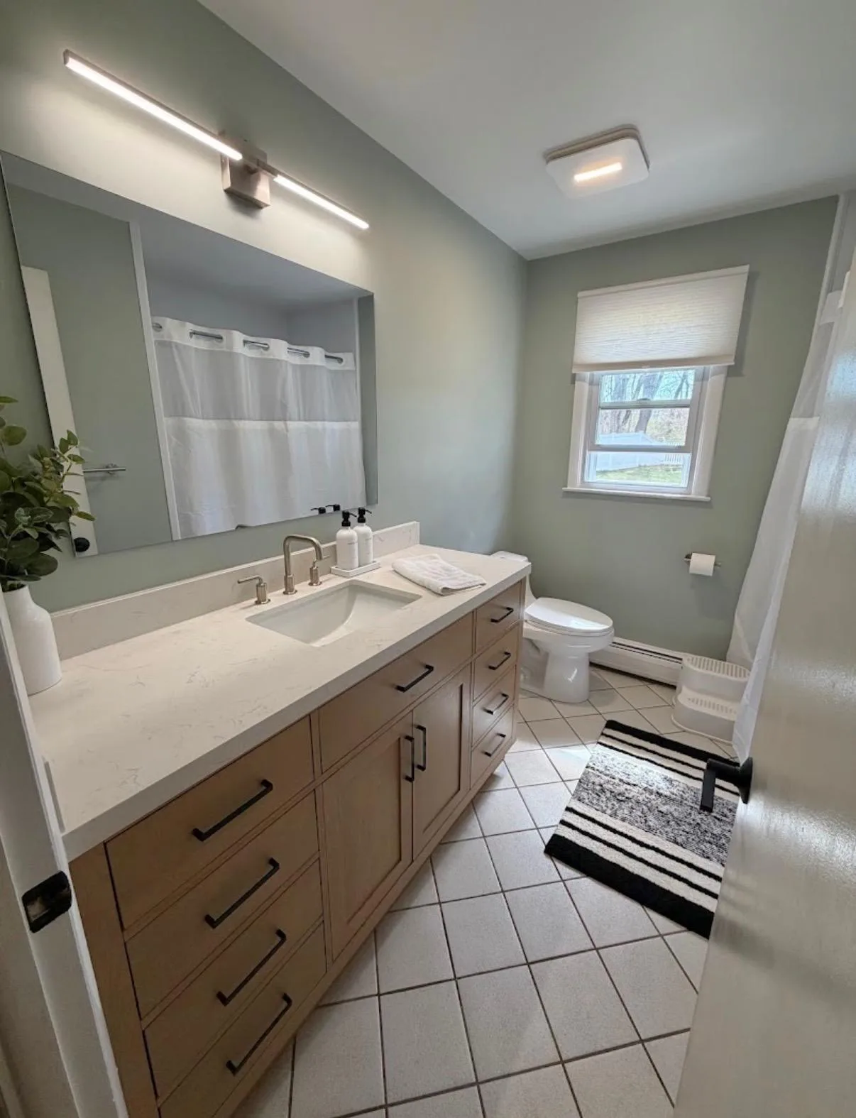 After the modern bathroom remodel, featuring updated sage green walls, a natural wood vanity with dark hardware, and sleek contemporary lighting.